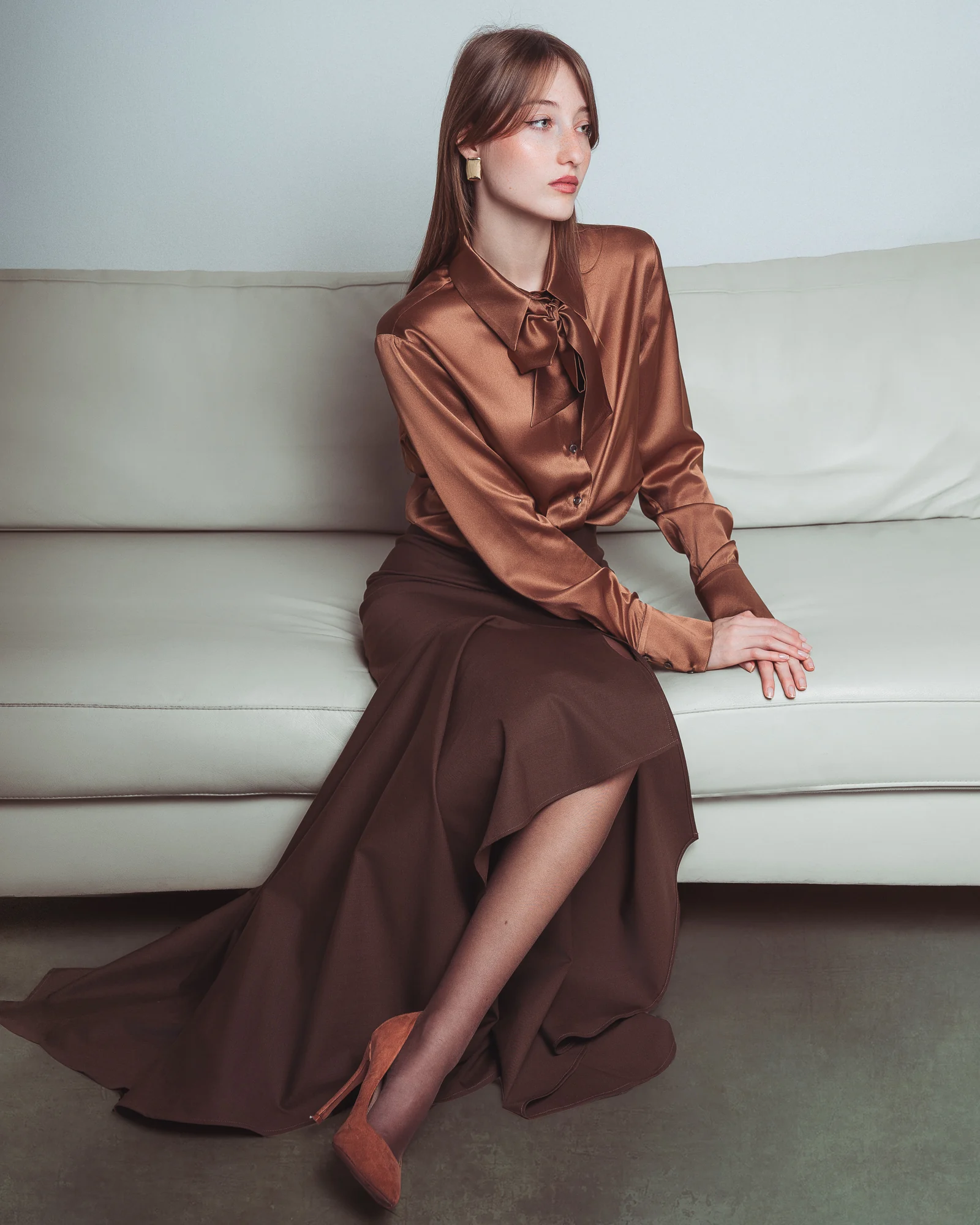 Incantevole Milano Fall Winter 2026–2027 – bronze blouse and flowing brown skirt, seated on a curved sofa